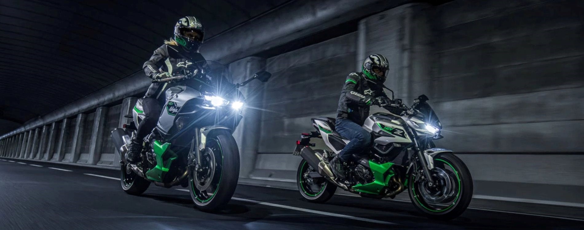 Zwei Motorradfahrer fahren nebeneinander auf den Kawasaki Hybrid-Motorrädern durch einen beleuchteten Tunnel. Beide tragen Schutzkleidung und Helme, während die Motorräder mit grünen Akzenten und eingeschaltetem LED-Licht auffallen. Die Szene vermittelt Geschwindigkeit und Dynamik in einer urbanen Umgebung.