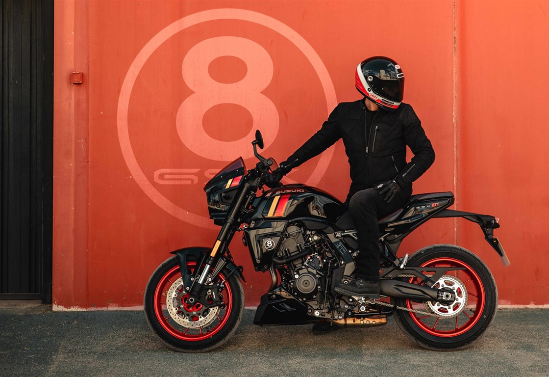 Fahrer in schwarzer Motorradkleidung sitzt auf einer Suzuki GSX-8TT im markanten Café-Racer-Design. Das Motorrad ist schwarz mit roten Felgen und sportlichen Farbakzenten auf dem Tank gestaltet. Im Hintergrund steht eine rote Wand mit großem 8-Ball-Logo.
