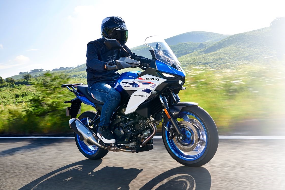 Eine Person mit schwarzem Motorradhelm fährt auf einer blau-weißen Suzuki SV-7GX über eine Landstraße vor grünen Hügeln